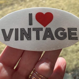 May include: Oval white sticker with the text "I ❤️ VINTAGE" in black, with a red heart symbol. The word "VINTAGE" is in a bold, striped font. The sticker is held by a hand with gold rings.