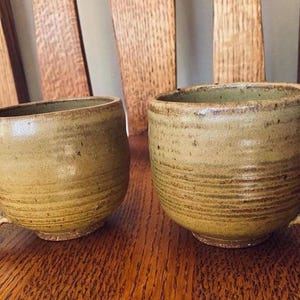 May include: Two ceramic mugs with a brown and tan glaze. The mugs have a rounded shape and a handle. The mugs are sitting on a wooden surface.