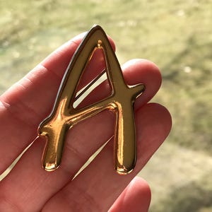 May include: A gold-colored letter "A" pin. The pin is in a cursive font and has a shiny, reflective surface. The pin is held in a hand against a blurred green background.