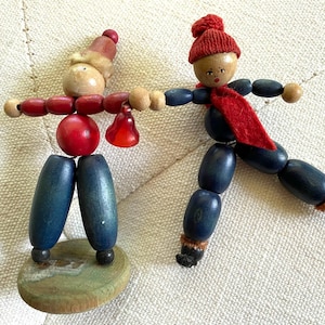 May include: Two wooden figurines. One has a red hat and scarf, the other a red and beige hat. Both have blue legs and arms. One holds a red bell. The figurines are on a cream-colored surface.