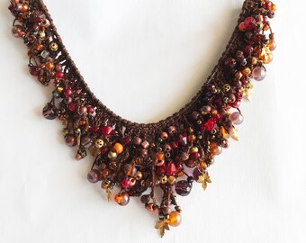 May include: A brown macrame necklace featuring a dense arrangement of multi-colored beads in shades of red, orange, amber, and bronze. The design includes small metallic leaf-shaped charms interspersed among faceted crystals and round glass beads.
