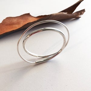 May include: A polished silver-tone bangle bracelet featuring a double-loop design with two overlapping circular bands of varying thickness. The metal has a smooth, reflective finish that catches the light, creating a clean and continuous circular form.
