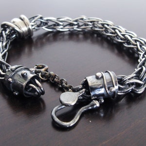 Könnte beinhalten: Ein silberfarbenes Kettenarmband mit einem geflochtenen Gliederkettendesign. Das Armband verfügt über zwei zylindrische Endkappen, die mit kleinen erhabenen Punkten verziert sind, sowie einen hakenförmigen Verschluss mit einer kurzen Kettenverlängerung.