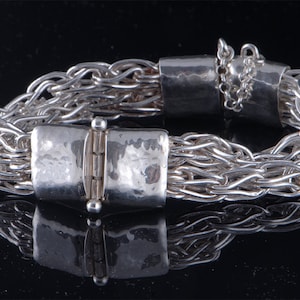 May include: A silver bracelet composed of multiple interwoven fine chains connected by a wide, hammered silver clasp. The clasp features a central hinge mechanism with a small pin closure and an attached safety chain for added security.