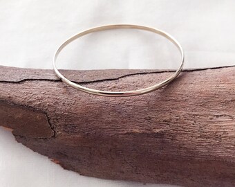 Può includere: Un bracciale rigido sottile in metallo color argento con una superficie liscia e arrotondata. Il bracciale circolare poggia su un pezzo di legno marrone scuro testurizzato, caratterizzato da crepe naturali e un bordo ruvido e irregolare.