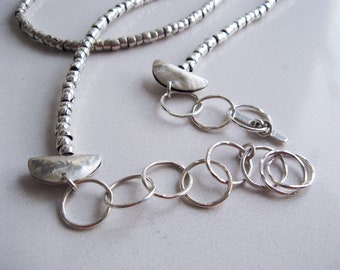 May include: A silver-toned necklace featuring a strand of small, textured cylindrical beads. The design includes two polished, semi-circular metal accents connected to a series of interlocking circular links that form an adjustable chain closure.