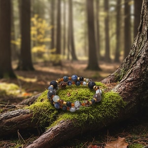 Puede incluir: Una pulsera de múltiples hilos con varias cuentas en tonos azules, marrones, negros y blancos, que descansa sobre un lecho de musgo verde. La pulsera se encuentra sobre un fondo borroso de bosque, con la luz del sol filtrándose a través de los árboles. La pulsera tiene una circunferencia de unos 18 cm.