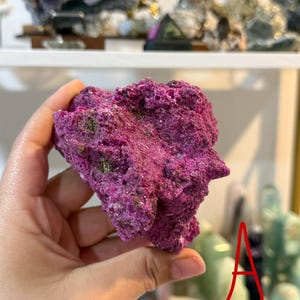Op de afbeelding: Een levendig, getextureerd, magenta-kleurig mineraalmonster. Het ruwe, poreuze oppervlak heeft hints van groen en wit. Het mineraal wordt in een hand gehouden, met andere exemplaren zichtbaar op een plank op de achtergrond. De letter "A" is rood geschreven.