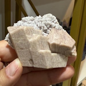 Puede incluir: Un espécimen mineral de color rosa claro y blanco. El espécimen tiene una estructura cristalina en capas con una superficie rugosa y texturizada. Los colores varían del rosa pálido al blanco, con una apariencia ligeramente translúcida. El mineral se sostiene en una mano.