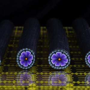 May include: Four cylindrical polymer clay canes arranged in a row, each featuring a vibrant purple flower pattern with a yellow centre and a teal-accented border on the end, set against a black background on a yellow measuring grid.