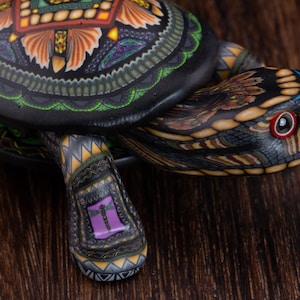 May include: A decorative turtle figurine featuring a black shell with intricate orange, green, and purple geometric patterns. The head and limbs are detailed with similar multi-coloured designs, including a small purple dragonfly motif on the leg.