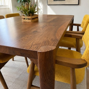 May include: A rectangular wooden dining table with a smooth brown grain surface and thick rounded legs, accompanied by several chairs featuring mustard yellow textured fabric seats and wooden frames, with a small wooden crate planter.
