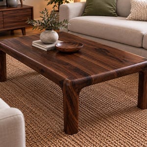 May include: A rectangular coffee table crafted from dark brown wood with a smooth, polished finish. The table features four thick, rounded legs and a solid tabletop surface, showcasing a prominent natural wood grain pattern throughout the piece.