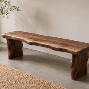 May include: A long wooden bench featuring a natural live edge top with a rich brown grain pattern. The bench is supported by two thick, sturdy slab legs that mirror the organic, uneven texture of the wood, showcasing a raw and solid construction.