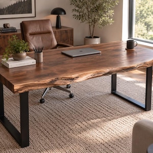 Peut inclure: Un bureau rectangulaire doté d'un plateau en bois massif à bord naturel avec un grain brun riche, soutenu par deux pieds en métal noir en forme de U. La surface du bureau supporte un ordinateur portable fermé, une petite plante et un livre.
