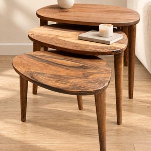May include: A set of three nesting tables crafted from brown wood with visible grain patterns. Each table features an oval-shaped top and three tapered legs. The tables are arranged in a cascading formation, with a white vase and a candle on top.