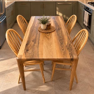 May include: A rectangular dining table made of natural wood with prominent dark grain patterns, featuring four matching spindle-back chairs. A round woven placemat sits in the centre, holding a small white vase with green foliage and salt shakers.