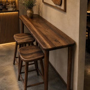 Puede incluir: Una mesa de bar alta y rectangular de madera con esquinas redondeadas y dos taburetes de bar a juego. El mobiliario está hecho de madera marrón oscura con una veta visible. Un jarrón de cerámica con follaje verde se encuentra sobre la mesa.