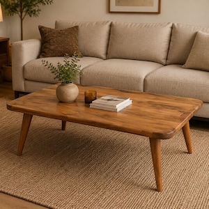 May include: A rectangular wooden coffee table with rounded corners and four tapered legs. The table surface features a natural wood grain finish and holds a small ceramic vase with green foliage, a glass candle holder, and two stacked books.