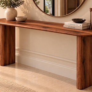 Peut inclure: Une console longue et &eacute;troite en bois brun fonc&eacute; avec un plateau aux bords arrondis et deux pieds &eacute;pais de forme cylindrique. La surface de la table supporte un vase en c&eacute;ramique, un petit bol, deux livres empil&eacute;s, un bol sombre et deux bougeoirs.