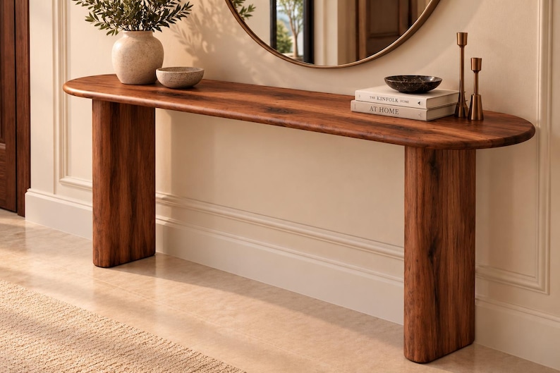 Peut inclure: Une console longue et &eacute;troite en bois brun fonc&eacute; avec un plateau aux bords arrondis et deux pieds &eacute;pais de forme cylindrique. La surface de la table supporte un vase en c&eacute;ramique, un petit bol, deux livres empil&eacute;s, un bol sombre et deux bougeoirs.