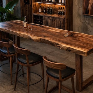 Op de afbeelding: Een lange rechthoekige bartafel met een massief houten blad met natuurlijke boomstamrand en donkerbruine nerf, ondersteund door stevige houten poten, vergezeld van vier bijpassende barkrukken met zwarte gestoffeerde zittingen en gebogen rugleuningen.