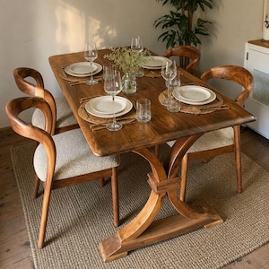 May include: A rectangular wooden dining table with a trestle base and four matching chairs featuring curved wooden frames and cream-colored textured fabric seats. The table is set with four white ceramic plates, clear wine glasses, and tumblers.