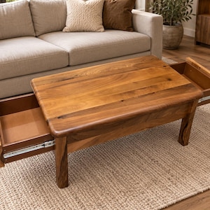 Op de afbeelding: Een rechthoekige houten salontafel met een natuurlijke bruine houtnerfafwerking, afgeronde hoeken en vier stevige poten. De tafel bevat twee lades die aan tegenovergestelde zijden zijn uitgetrokken, wat gladde binnenoppervlakken en metalen geleiders toont.