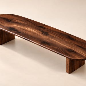 May include: A low, elongated bench crafted from dark brown wood with a prominent natural grain pattern. The piece features a smooth, oval-shaped top surface supported by two thick, solid wooden legs positioned at each end of the structure.