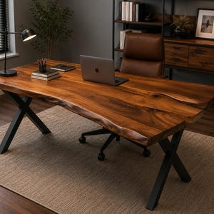 Puede incluir: Un escritorio grande de madera con un acabado de borde natural y una veta marrón rica, sostenido por patas de metal negro en forma de X. Un portátil plateado, una lámpara de escritorio negra y libros están colocados sobre la superficie.