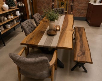 Pode incluir: Uma mesa de jantar de madeira com borda natural, um caminho de mesa bege, uma tigela de madeira, um vaso com folhagens e uma vela. O conjunto inclui três cadeiras estofadas cinzas com capitonê e um banco de madeira a condizer com pernas pretas.