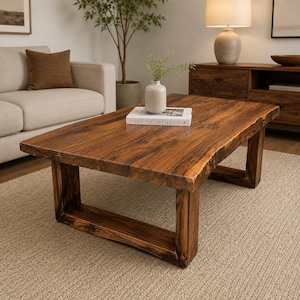 May include: A rectangular coffee table made of dark brown wood with a natural live edge finish. The table features a thick, solid tabletop supported by two sturdy, U-shaped wooden legs. A white book titled LIVE BEAUTIFULLY sits on top.