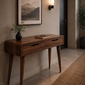Peut inclure: Une console en bois foncé aux bords arrondis, dotée de quatre pieds fuselés et de deux tiroirs avec poignées horizontales intégrées. Un petit vase noir avec du feuillage vert et un carnet fermé reposent sur la surface en bois poli.