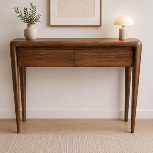Può includere: Un tavolo console in legno scuro con due cassetti, caratterizzato da bordi arrotondati e gambe affusolate. Sopra sono posizionati un vaso in ceramica chiara con fogliame verde e una piccola lampada da tavolo con paralume bianco e base in pietra.