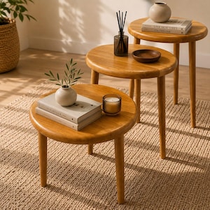 Puede incluir: Un conjunto de tres mesas nido redondas de madera con superficies lisas y patas cónicas. Las mesas están dispuestas en formación escalonada, cada una con objetos decorativos que incluyen libros, un pequeño jarrón con vegetación y una vela.