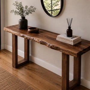 Peut inclure: Une longue console dotée d'un plateau en bois brun foncé à bord naturel et de deux pieds rectangulaires robustes. La surface de la table supporte un vase sombre avec de la verdure, un petit bol en bois, deux livres et un diffuseur à roseaux.