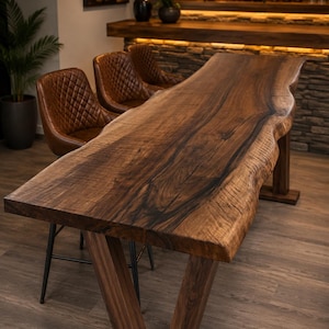 Puede incluir: Una mesa de bar de madera de nogal con borde natural, veteado visible y acabado marrón oscuro, sostenida por dos patas de madera en forma de V. Tres taburetes de bar de cuero marrón con costuras en forma de diamante se sitúan junto a la mesa.
