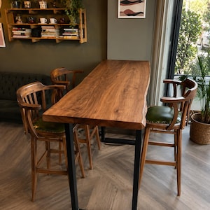 Peut inclure: Une table de bar rectangulaire haute avec un plateau en bois naturel à bord brut et des pieds en U en métal noir, accompagnée de trois tabourets de bar en bois avec dossiers incurvés et sièges rembourrés vert olive à clous décoratifs.