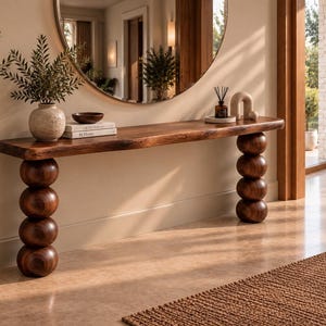 Op de afbeelding: Een lange houten sidetable met een natuurlijk boomstam-tafelblad en twee poten bestaande uit stapels van vier grote, gladde, donkerbruine houten bollen. Op het tafelblad staan een keramische vaas, een kleine kom en twee gestapelde boeken.
