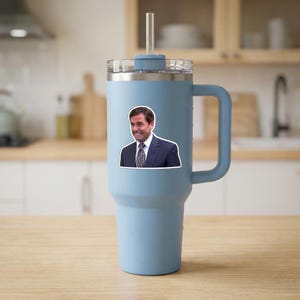 May include: A light blue insulated tumbler with a handle and a clear straw. A sticker of a man in a suit is affixed to the tumbler. The tumbler is sitting on a wooden surface.