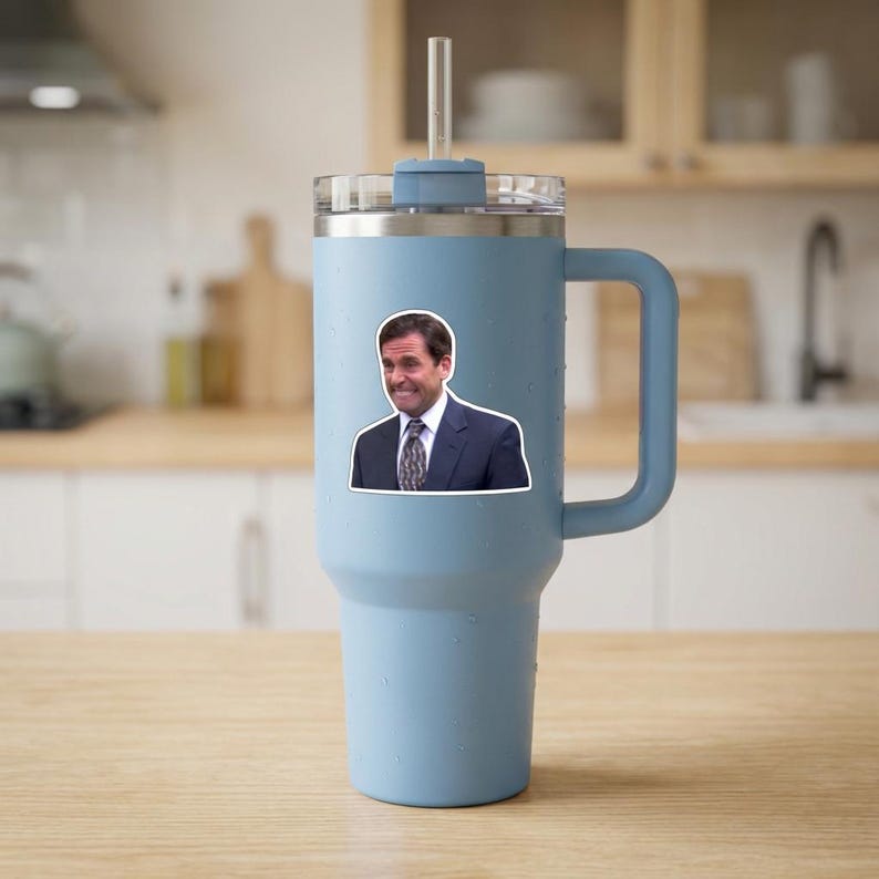 May include: A light blue insulated tumbler with a handle and a clear straw. A sticker of a man in a suit is affixed to the tumbler. The tumbler is sitting on a wooden surface.