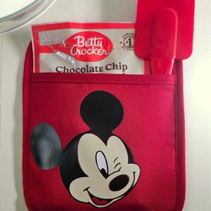 May include: Red oven mitt with a Mickey Mouse graphic, holding a Betty Crocker Chocolate Chip mix packet and two red spatulas. The mitt has a pocket and a loop for hanging. A clear glass bowl is in the background.