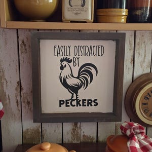 May include: A framed sign with the text "EASILY DISTRACTED BY PECKERS" and a black rooster illustration. The sign is in a dark brown frame and hangs on a wooden wall. Other kitchen items are visible on shelves.