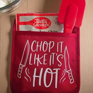 May include: Red oven mitt with the text "CHOP IT LIKE IT'S HOT" in white. A red spatula and a Betty Crocker baking mix are in the pocket. A bowl of white balls is in the background. Kitchenware.