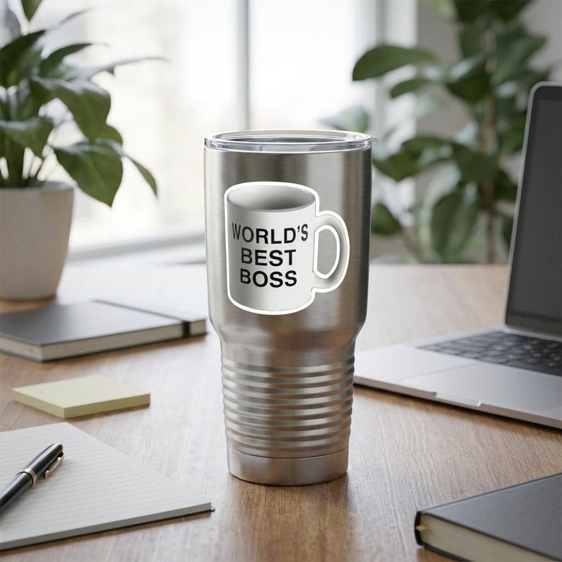 May include: A stainless steel tumbler with a white sticker of a coffee mug that reads "WORLD'S BEST BOSS" in black text. The tumbler is cylindrical with a ridged design and a silver finish. The background includes a laptop, notepad, and a plant.