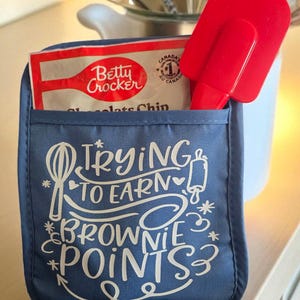 Puede incluir: Un guante de horno azul con el texto "Trying to Earn Brownie Points" en escritura blanca. Una espátula roja y un paquete de mezcla para galletas con chispas de chocolate Betty Crocker están en el bolsillo del guante.