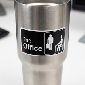 May include: A stainless steel tumbler with a black and white sticker. The sticker features the text "The Office" and a graphic of two stick figures, one standing with a briefcase and the other seated at a desk. The tumbler is cylindrical.