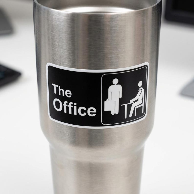 May include: A stainless steel tumbler with a black and white sticker. The sticker features the text "The Office" and a graphic of two stick figures, one standing with a briefcase and the other seated at a desk. The tumbler is cylindrical.