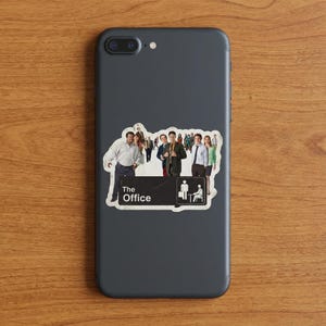 May include: A sticker featuring a group of people, with the text "The Office" and a small graphic of a person with a suitcase and a person sitting in a chair. The sticker is on a dark gray phone.