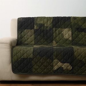May include: A quilted sofa cover in a camouflage pattern, featuring shades of green and black. The cover is made of a diamond-stitched fabric and is draped over the arm and seat of a beige sofa. The cover's edges are finished with a dark green trim.